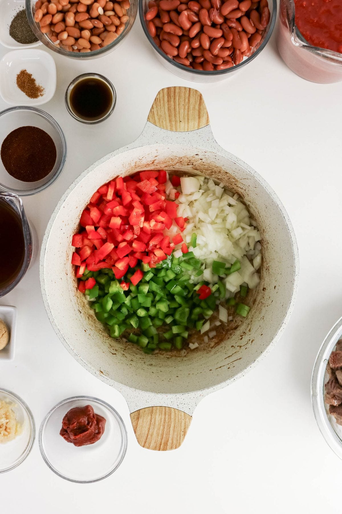 A pot with chopped tomatoes, onions, and green bell peppers, surrounded by bowls of beans, spices, tomato paste, broth, garlic, and other chili ingredients on a white surface.