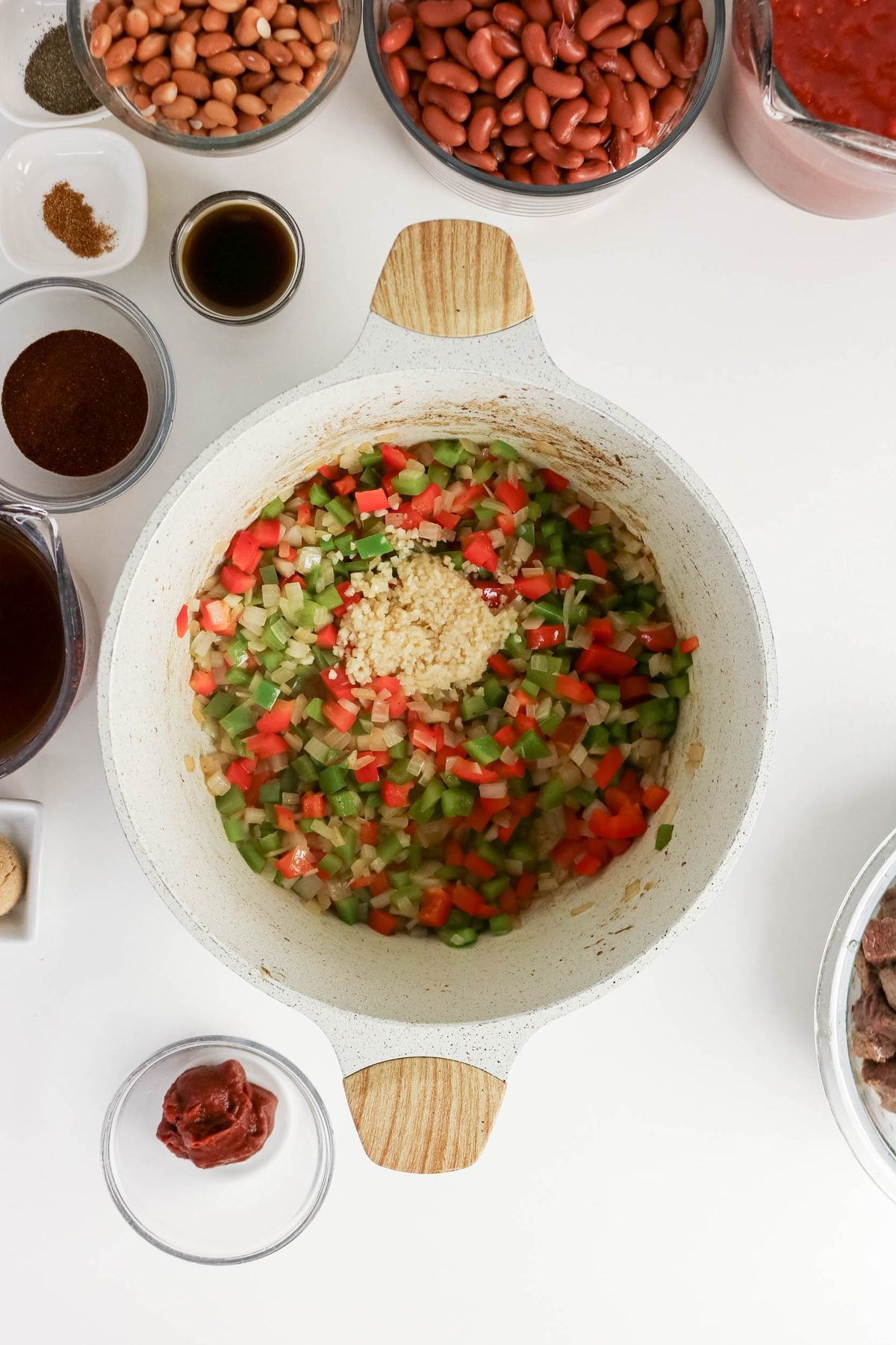 White pot with chopped onions, celery, and red bell peppers cooking, minced garlic in the center; surrounded by bowls of beans, spices, tomato paste, broth, and sauce on a white surface.