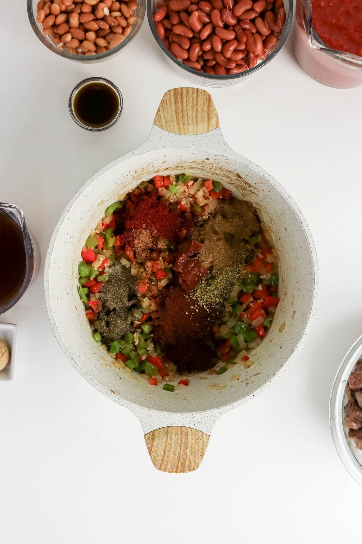 White pot with chopped onions, red and green bell peppers, and various spices on top, surrounded by bowls of beans, broth, tomato sauce, Worcestershire sauce, and meat on a white surface.