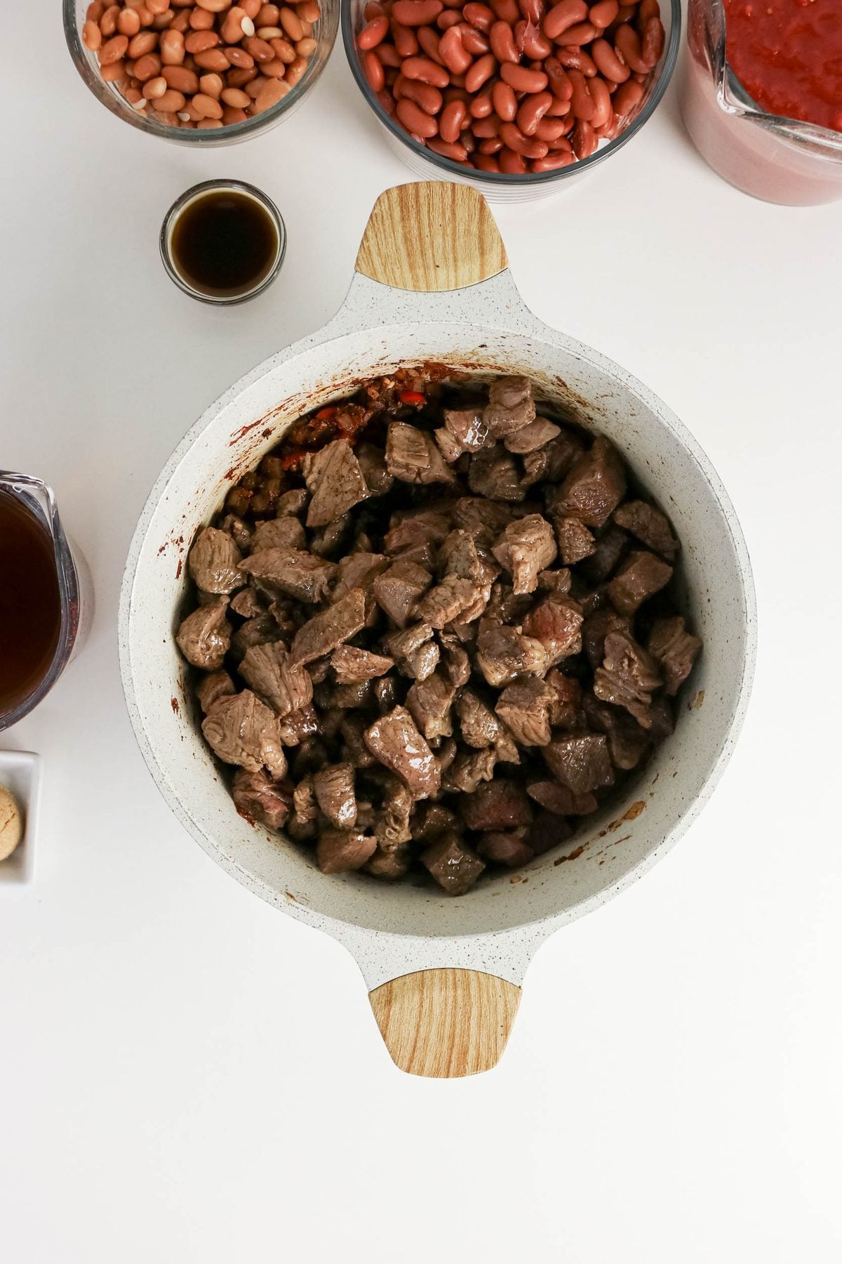 A white pot filled with cooked beef cubes sits on a white surface, surrounded by bowls of beans, tomato sauce, broth, and a small dish of sauce.