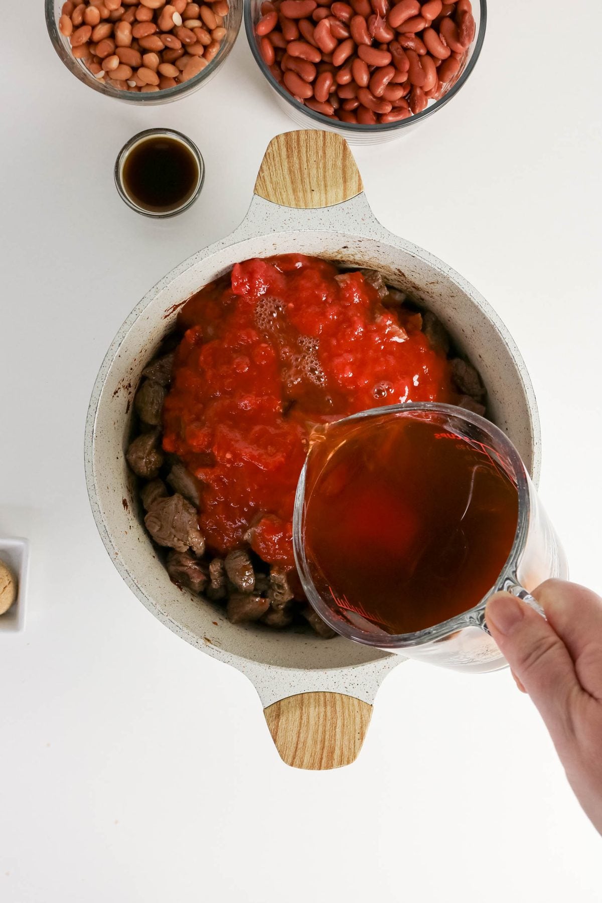 A hand pours broth into a pot containing beef and tomato sauce, with bowls of beans and a small dish of seasoning nearby on a white surface.