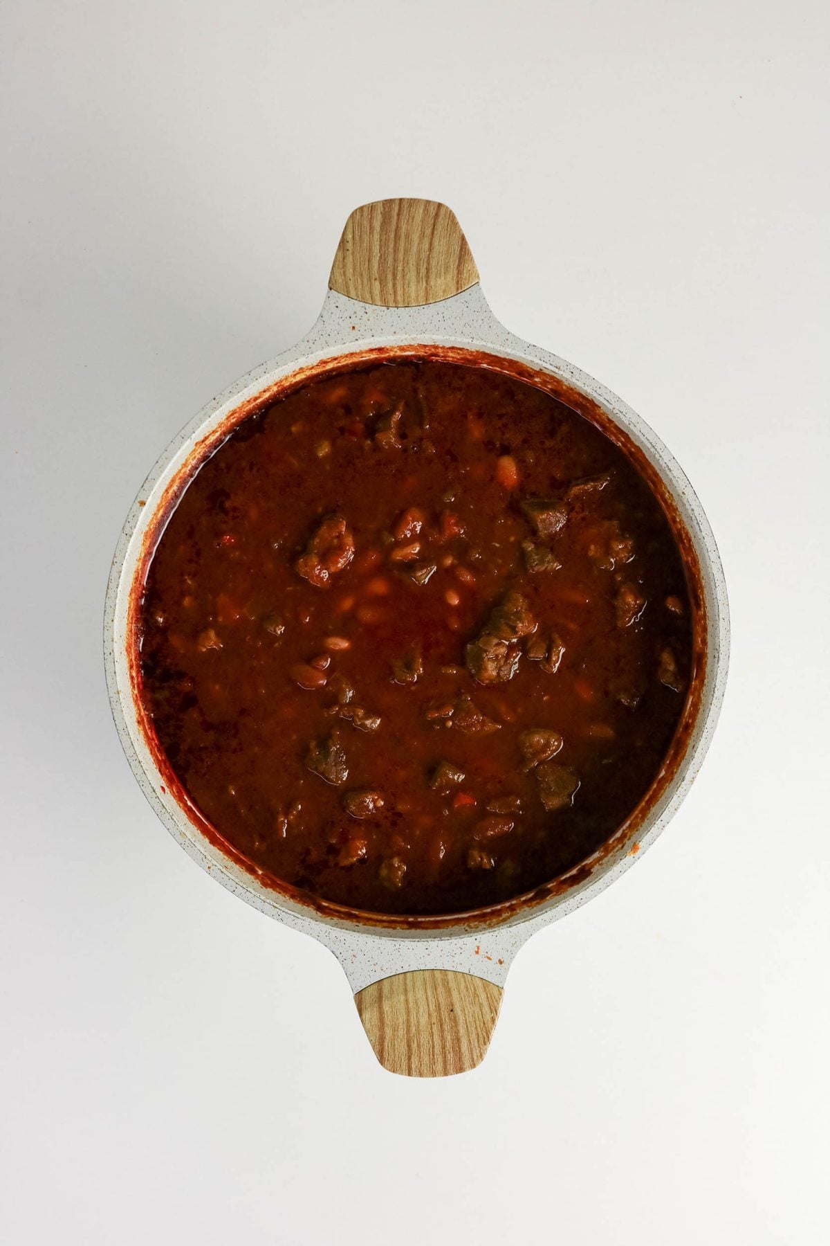 A ceramic pot filled with chunky beef and bean chili in red sauce, photographed from above on a white background.