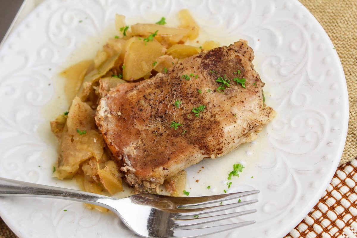 A serving of crockpot apple pork chops on a white plate with tender cooked apple slices and fresh parsley