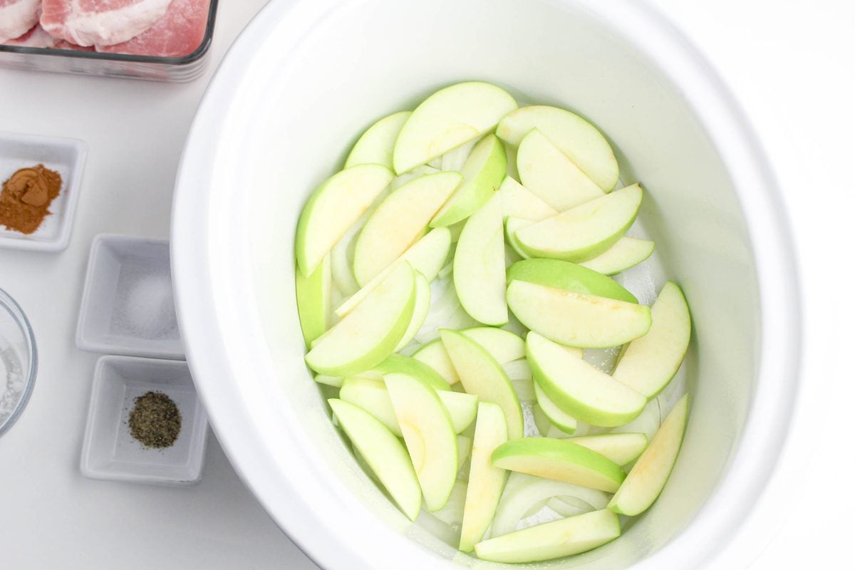 Step 1 for crockpot apple pork chops: layering sliced green apples in the bottom of a white slow cooker
