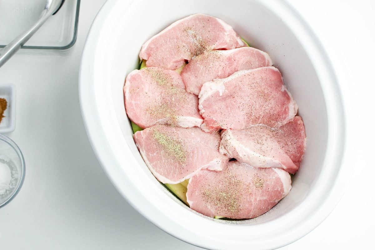 Step 2 for crockpot apple pork chops: placing raw seasoned pork chops over the apple slices in the slow cooker
