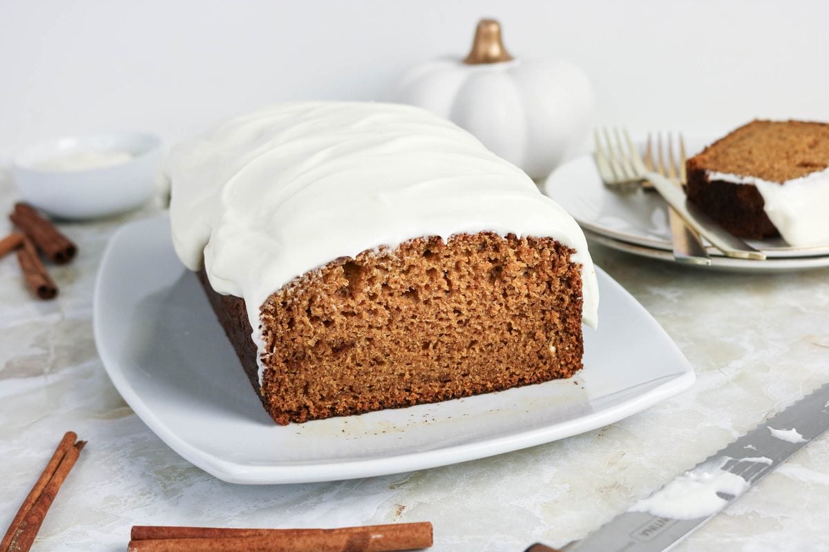 A loaf of spiced bread with white frosting is displayed on a white serving plate, with a slice cut and plated nearby. Cinnamon sticks and utensils are also visible.