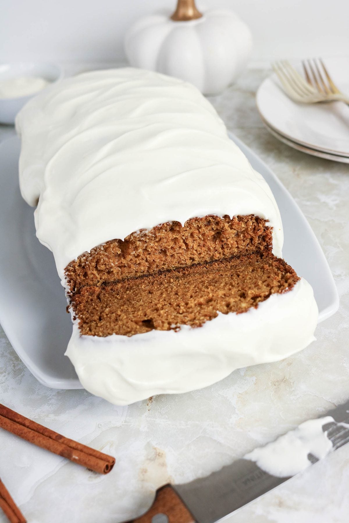 A loaf of brown spice cake covered in white icing, with one slice cut, is on a white plate beside a knife, plates, and forks. Cinnamon sticks and a small pumpkin are nearby.