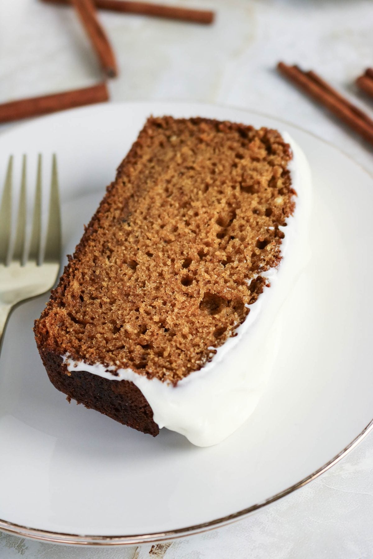A slice of brown spice cake with white icing sits on a white plate next to a fork, with cinnamon sticks in the background.