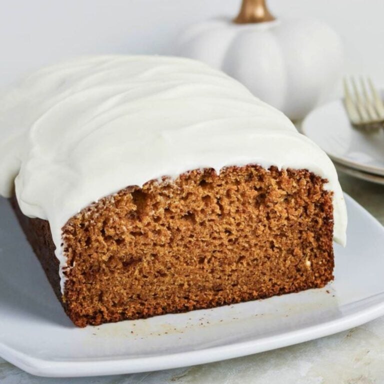 A loaf of pumpkin bread with white icing on top sits on a white plate, with a few slices cut. A white pumpkin and plates with forks are in the background.