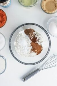 A glass bowl with flour, baking powder, cinnamon, nutmeg, and salt on a white surface, surrounded by bowls of sugar, eggs, milk, oil, vanilla, brown sugar, and a whisk.