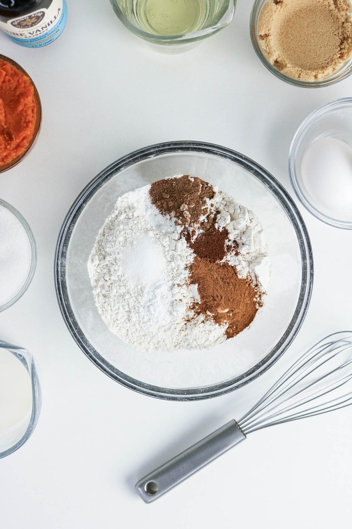A glass bowl with flour, baking powder, cinnamon, nutmeg, and salt on a white surface, surrounded by bowls of sugar, eggs, milk, oil, vanilla, brown sugar, and a whisk.