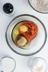 A glass bowl with eggs, canned pumpkin, brown sugar, white sugar, and oil, surrounded by a milk jug, a bottle of vanilla, flour, and eggs on a white surface.