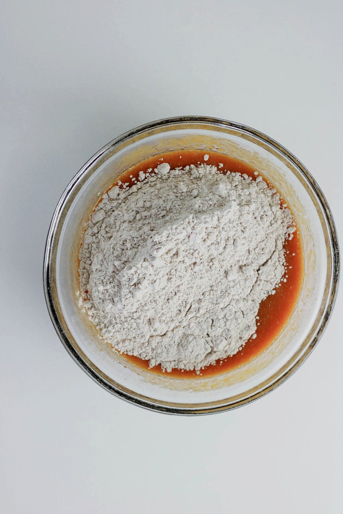 A glass bowl containing a mound of flour on top of a brownish liquid mixture, viewed from above on a white surface.