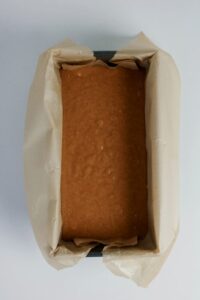 A rectangular loaf pan lined with parchment paper, filled with unbaked brown cake or bread batter, viewed from above.