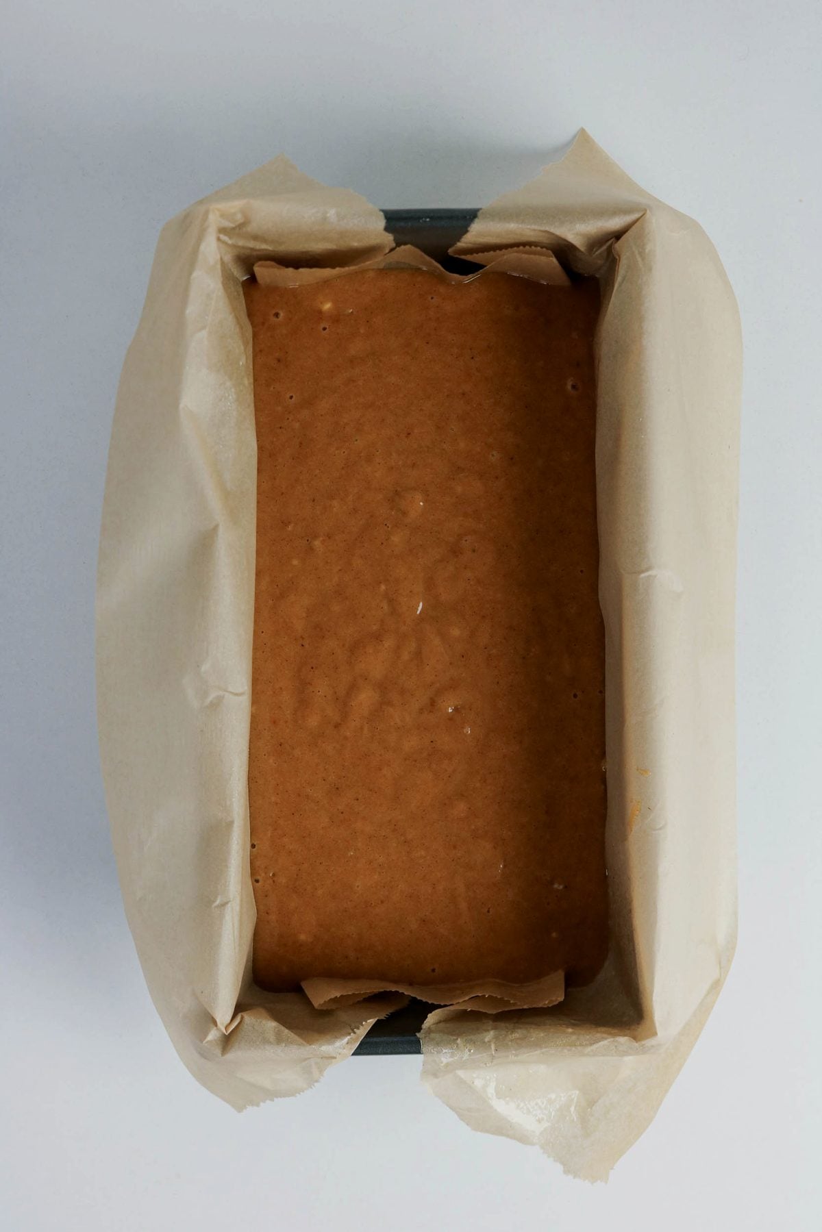 A rectangular loaf pan lined with parchment paper, filled with unbaked brown cake or bread batter, viewed from above.