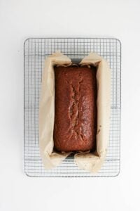 A loaf of banana bread in a parchment-lined pan sits on a metal cooling rack against a white background.