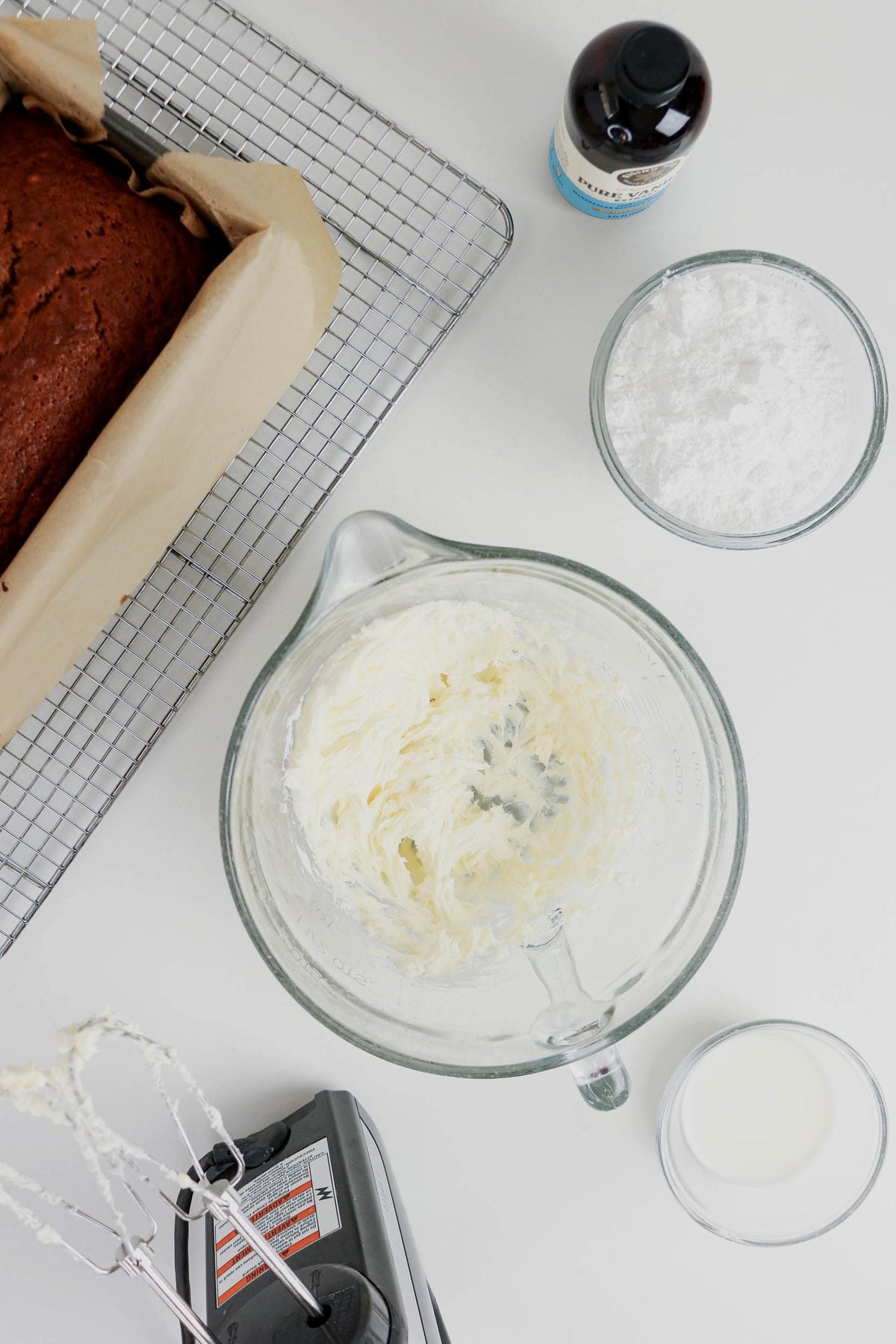 A glass bowl with creamed butter, a bowl of powdered sugar, milk, vanilla extract, a loaf cake on a rack, and a hand mixer on a white surface.