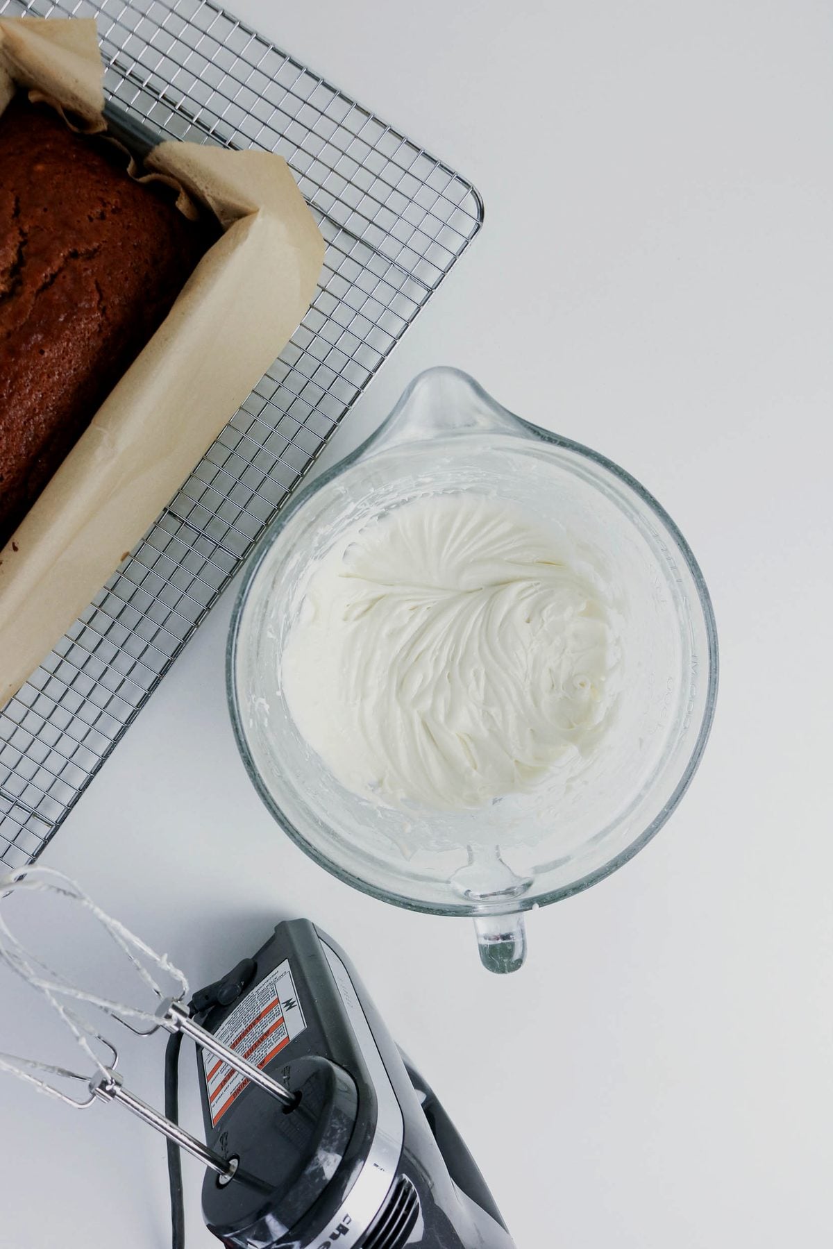 A glass bowl of whipped cream sits next to an electric mixer and a loaf of baked bread on a cooling rack, all on a white surface.