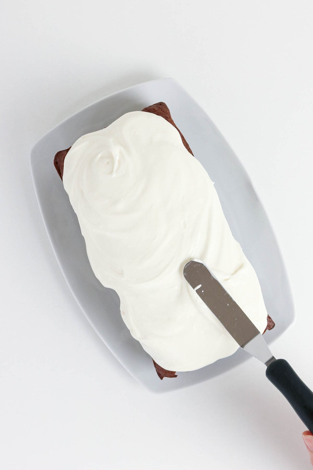 A rectangular cake on a white plate is being frosted with white icing using a metal spatula.