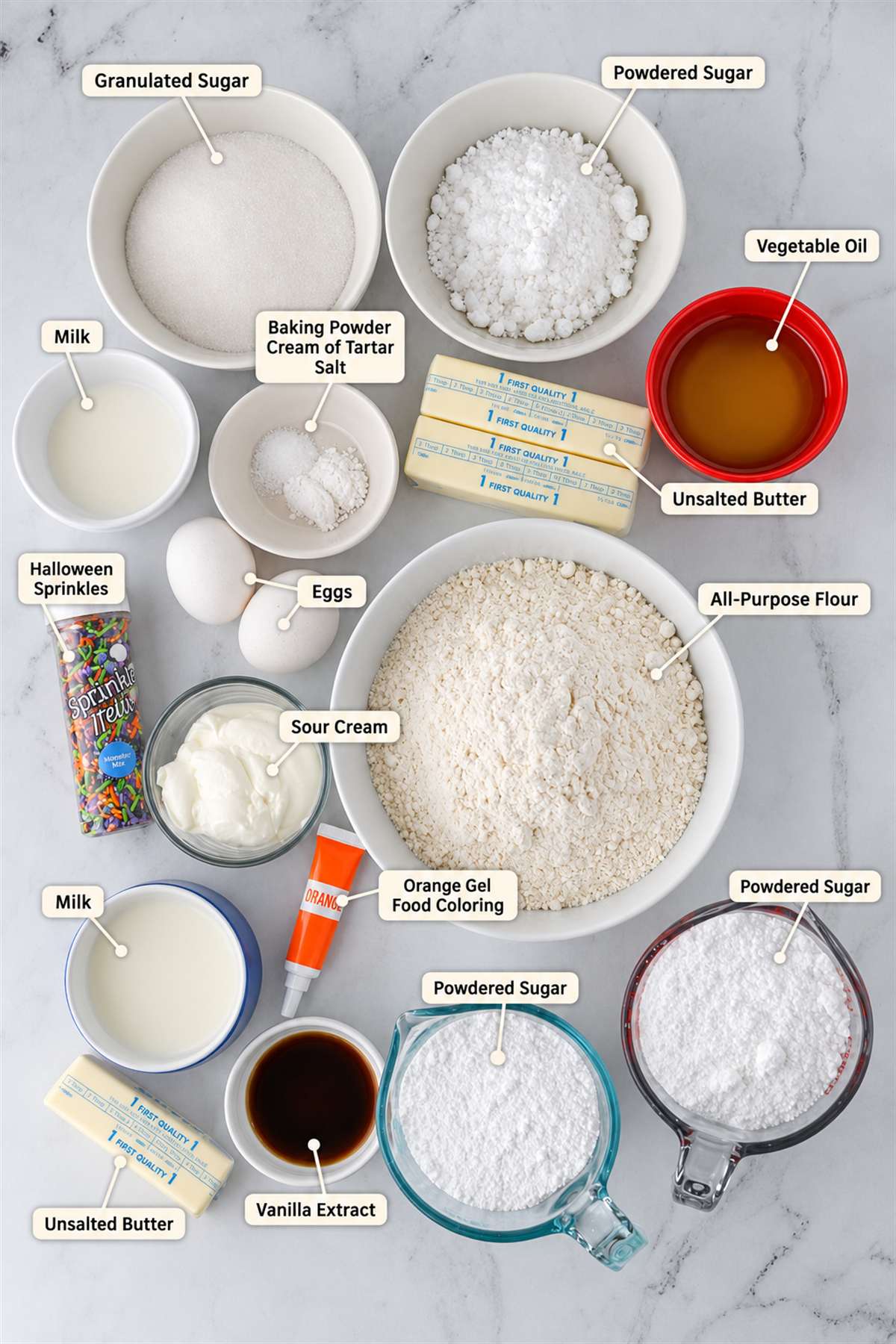 Baking ingredients arranged on a counter, including flour, sugars, eggs, sour cream, butter, oil, milk, vanilla, baking powder, food coloring, and Halloween sprinkles, each labeled.