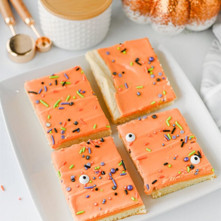 Four rectangular sugar cookie bars with orange frosting, sprinkles, and candy eyes are arranged on a white plate.
