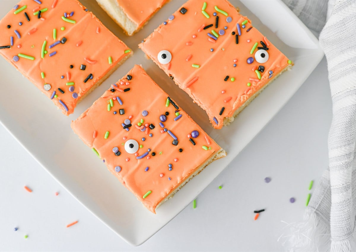 Four rectangular sugar cookies with orange icing and colorful Halloween-themed sprinkles are arranged on a white plate. Some sprinkles are scattered on the table.