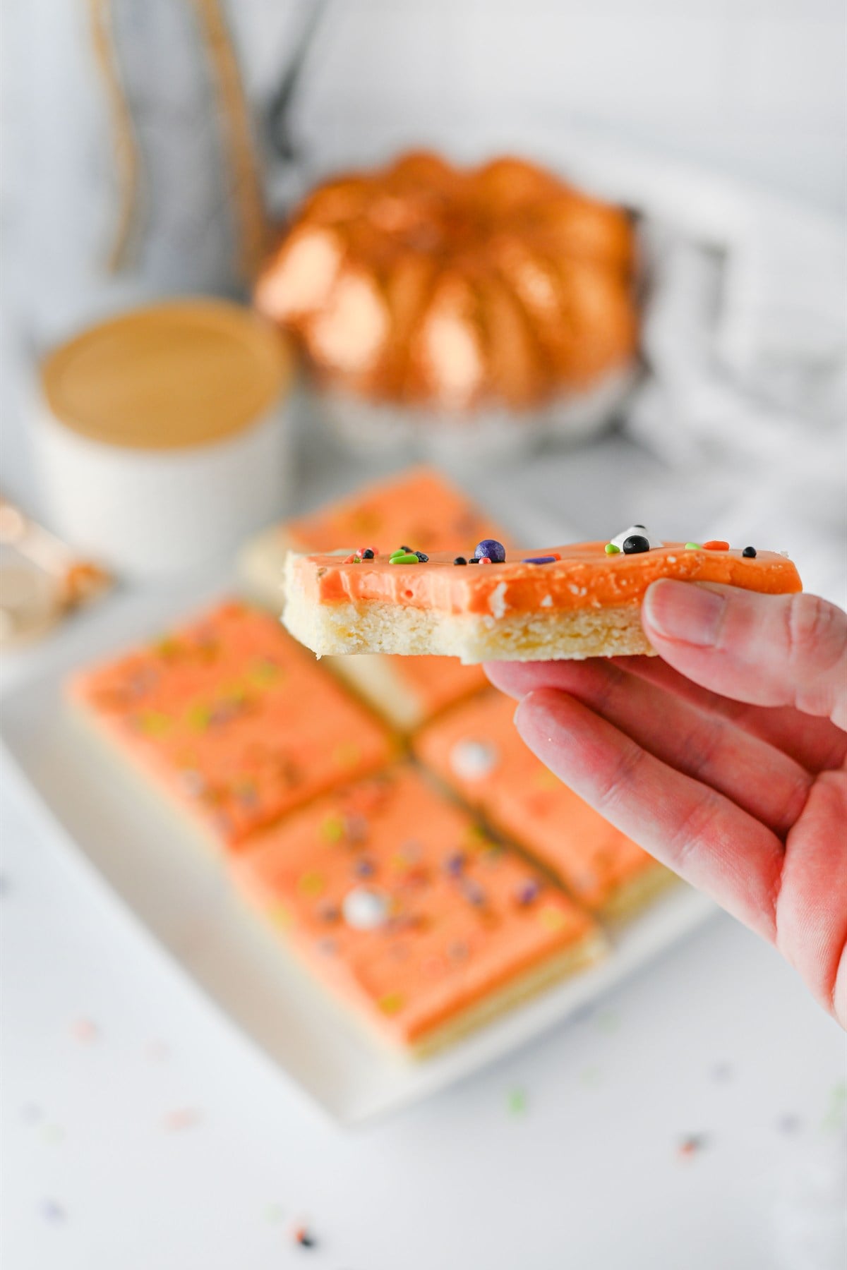 A hand holds a square cookie bar with orange frosting and sprinkles; more bars and a drink are on the table in the background.