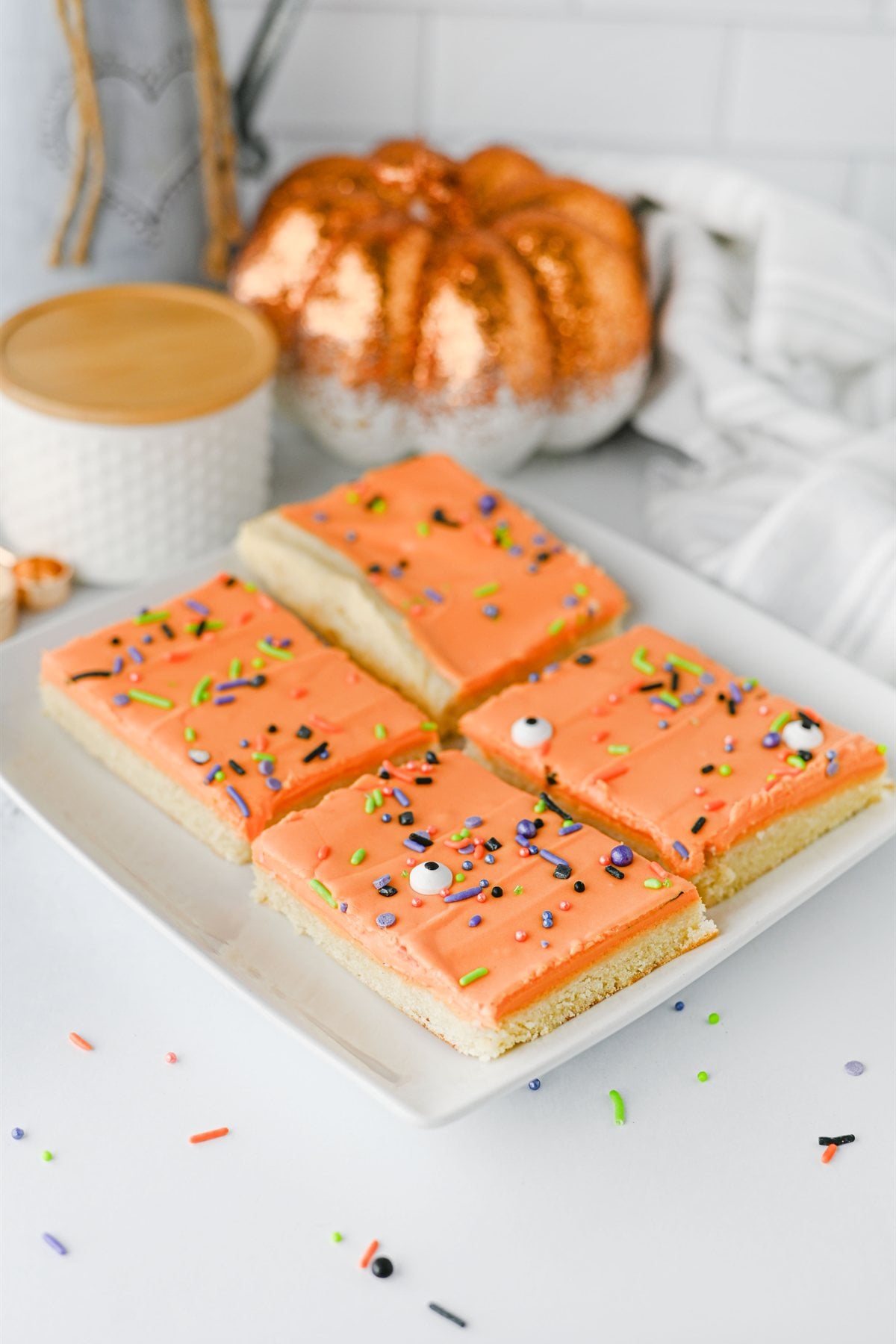 Four rectangular sugar cookie bars with orange frosting and Halloween-themed sprinkles on a white plate, with a decorative pumpkin in the background.