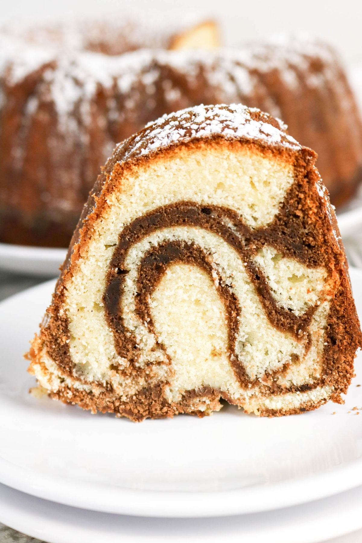 A slice of marble cake with swirled chocolate and vanilla layers, topped with powdered sugar, sits on a white plate.