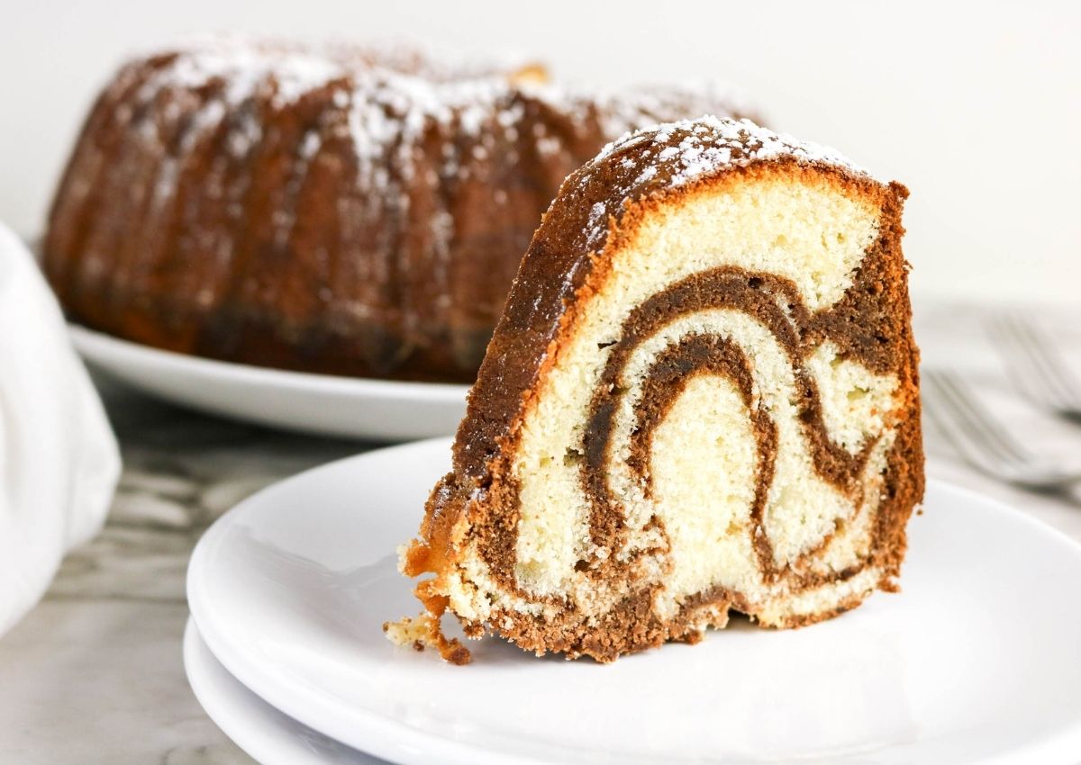 A slice of marble bundt cake with powdered sugar on top sits on a white plate in front of the whole cake.