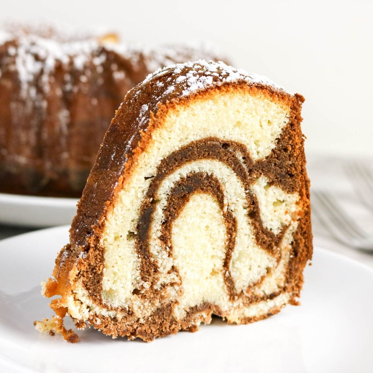 A slice of marble bundt cake with a swirl pattern of vanilla and chocolate, dusted with powdered sugar, sits on a white plate.