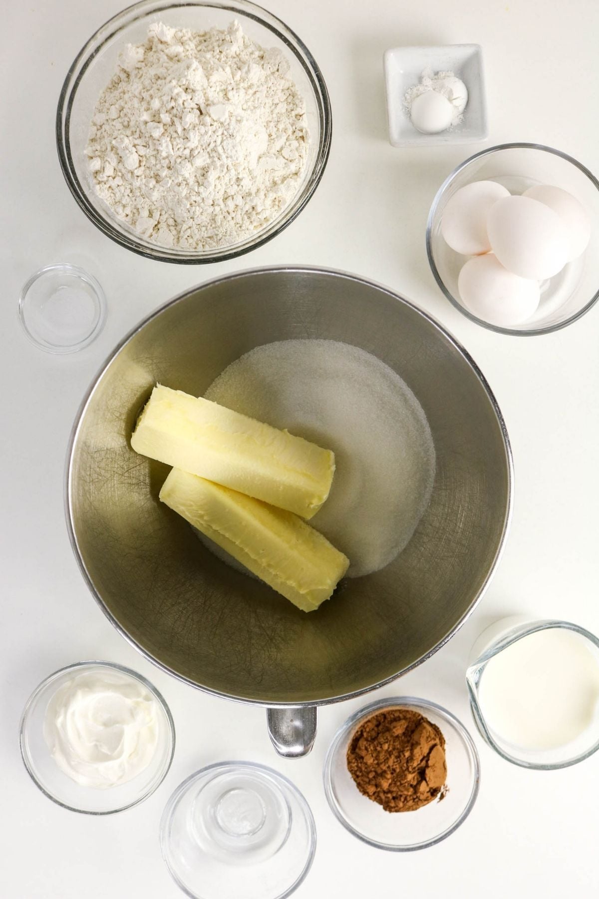 Bowls containing flour, baking powder, eggs, vanilla, milk, sour cream, cocoa powder, and a mixer bowl with butter and sugar, arranged on a white surface.