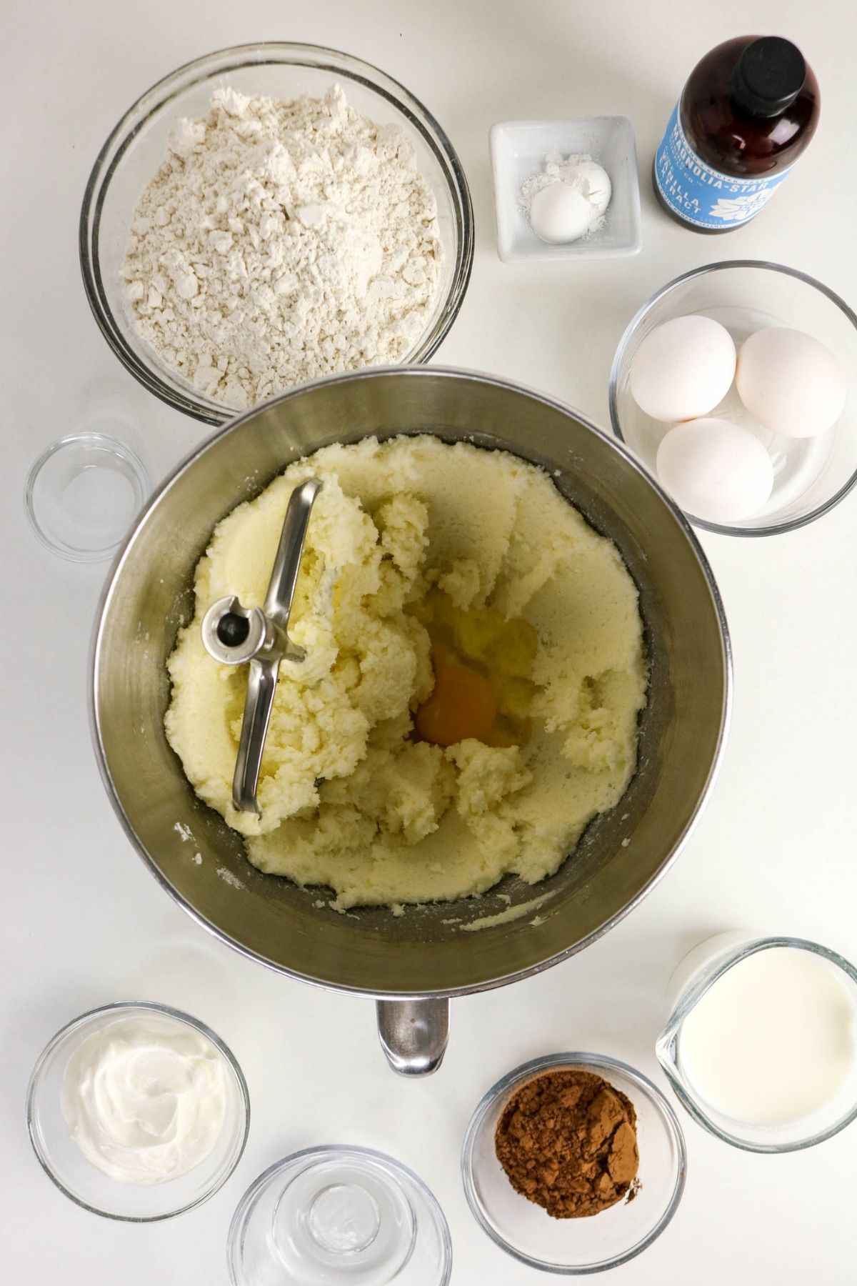 A mixing bowl with creamed butter and sugar, surrounded by bowls of flour, eggs, cocoa powder, sour cream, milk, salt, and vanilla extract on a white surface.
