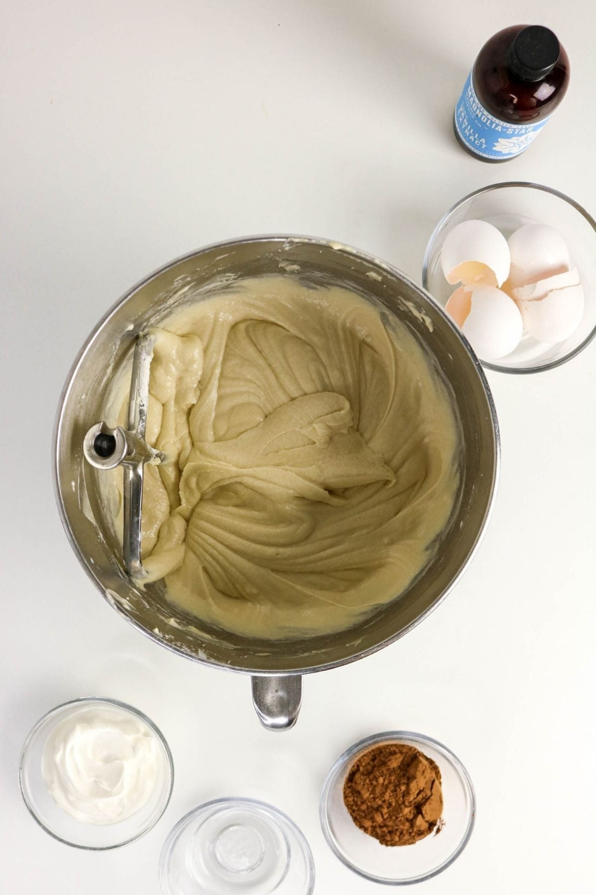 A mixing bowl with cake batter and a beater, surrounded by eggs, a bottle of vanilla, cocoa powder, and a bowl of cream on a white surface.