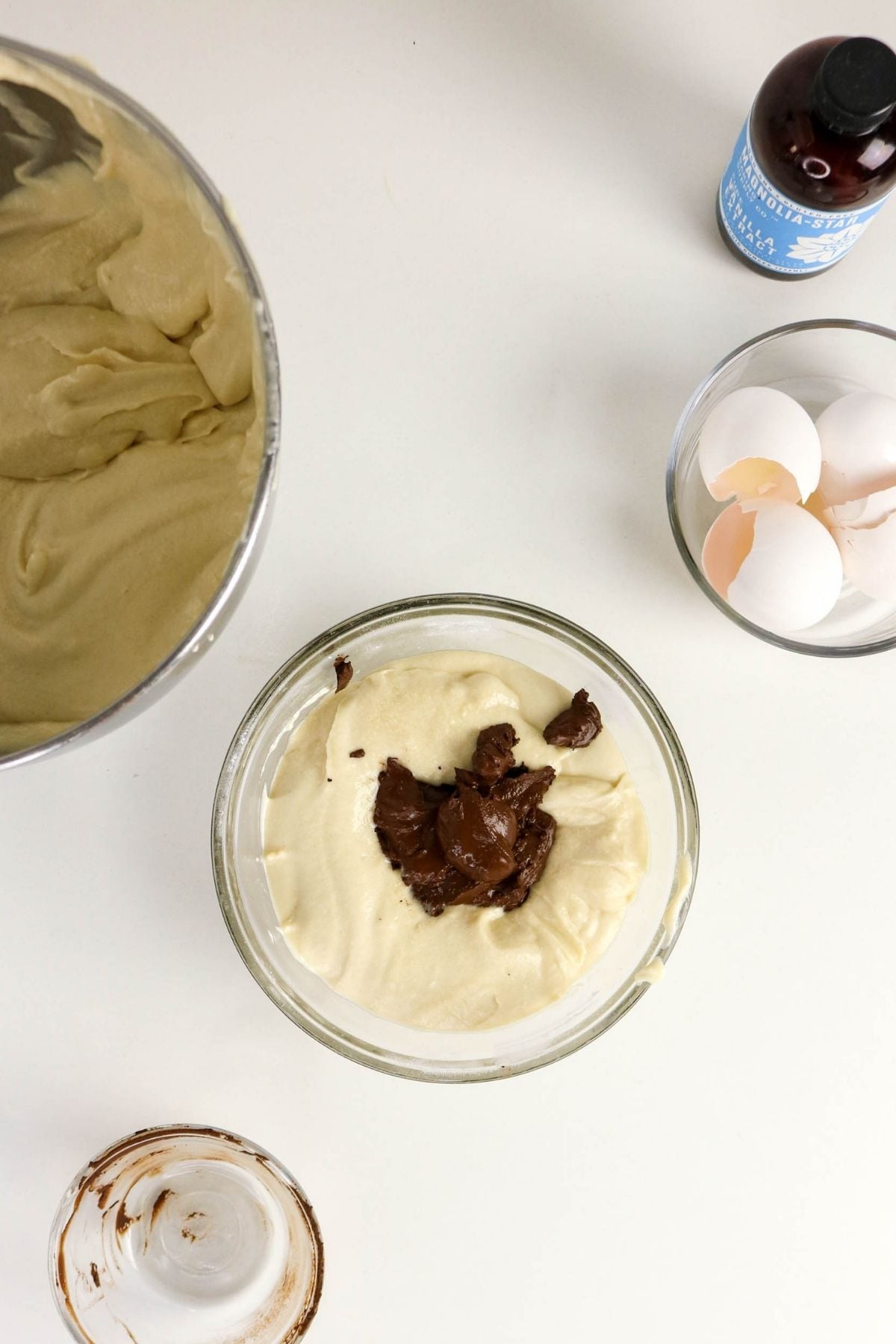 A glass bowl of cake batter with chocolate being folded in, next to a bowl of eggshells, a bottle of vanilla extract, and a mixing bowl on a white surface.