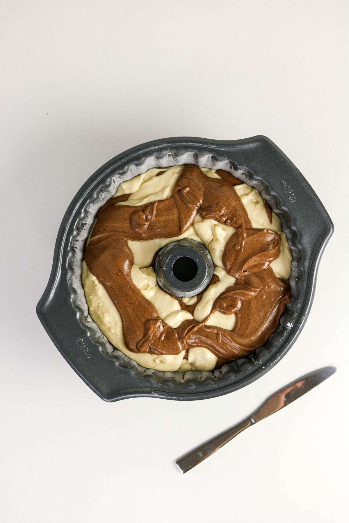 A bundt pan filled with swirled chocolate and vanilla cake batter sits on a white surface beside a butter knife.