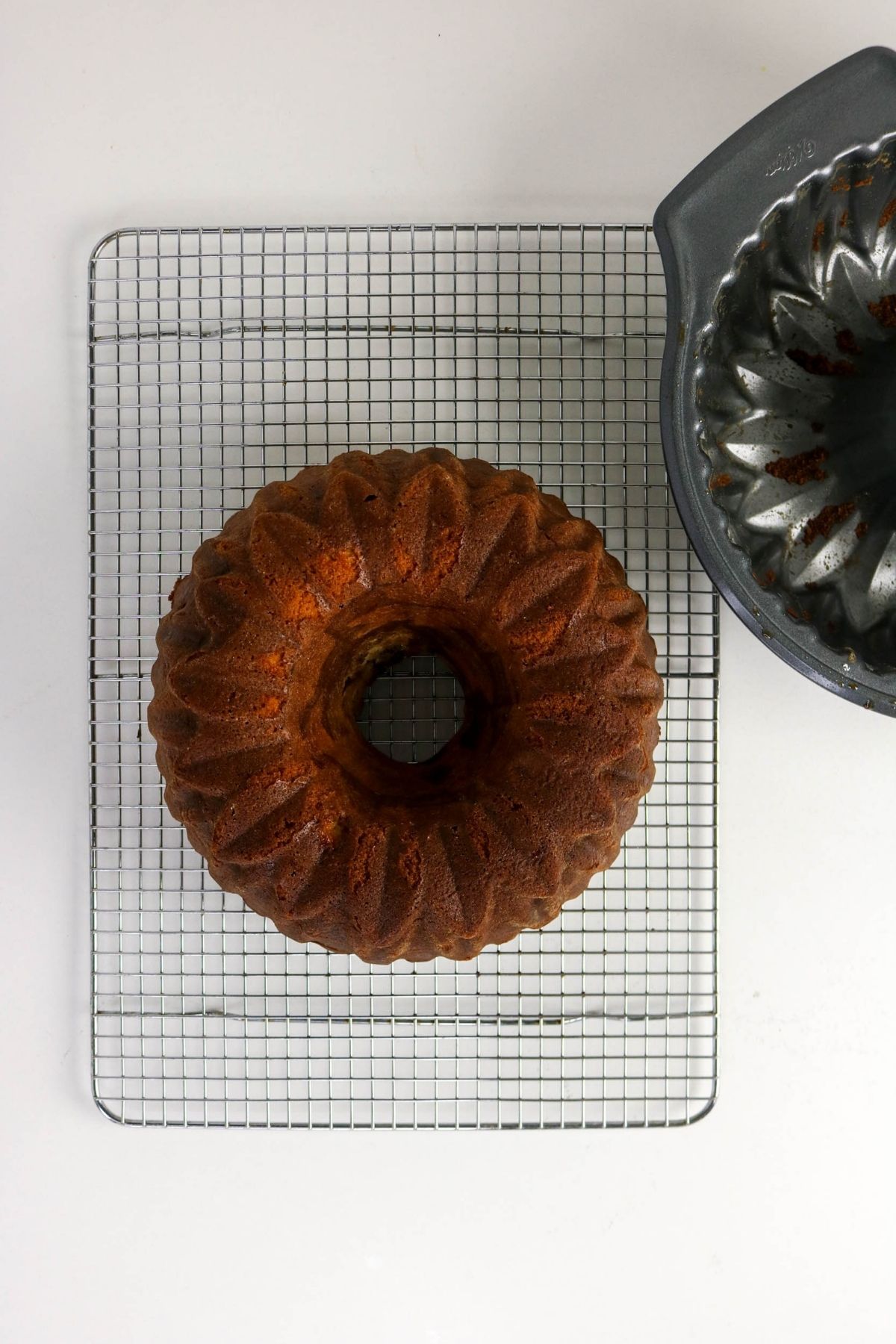 A round bundt cake cools on a wire rack next to an empty bundt cake pan, all placed on a white surface.