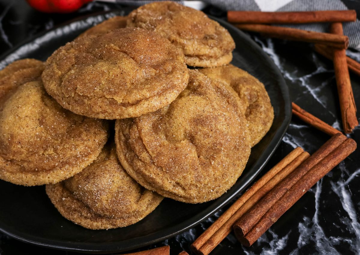 A plate of golden-brown cookies coated in cinnamon sugar, surrounded by cinnamon sticks on a black marble surface.