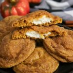 A plate of round, cinnamon-dusted cookies with one cookie broken in half, revealing a creamy white filling inside.