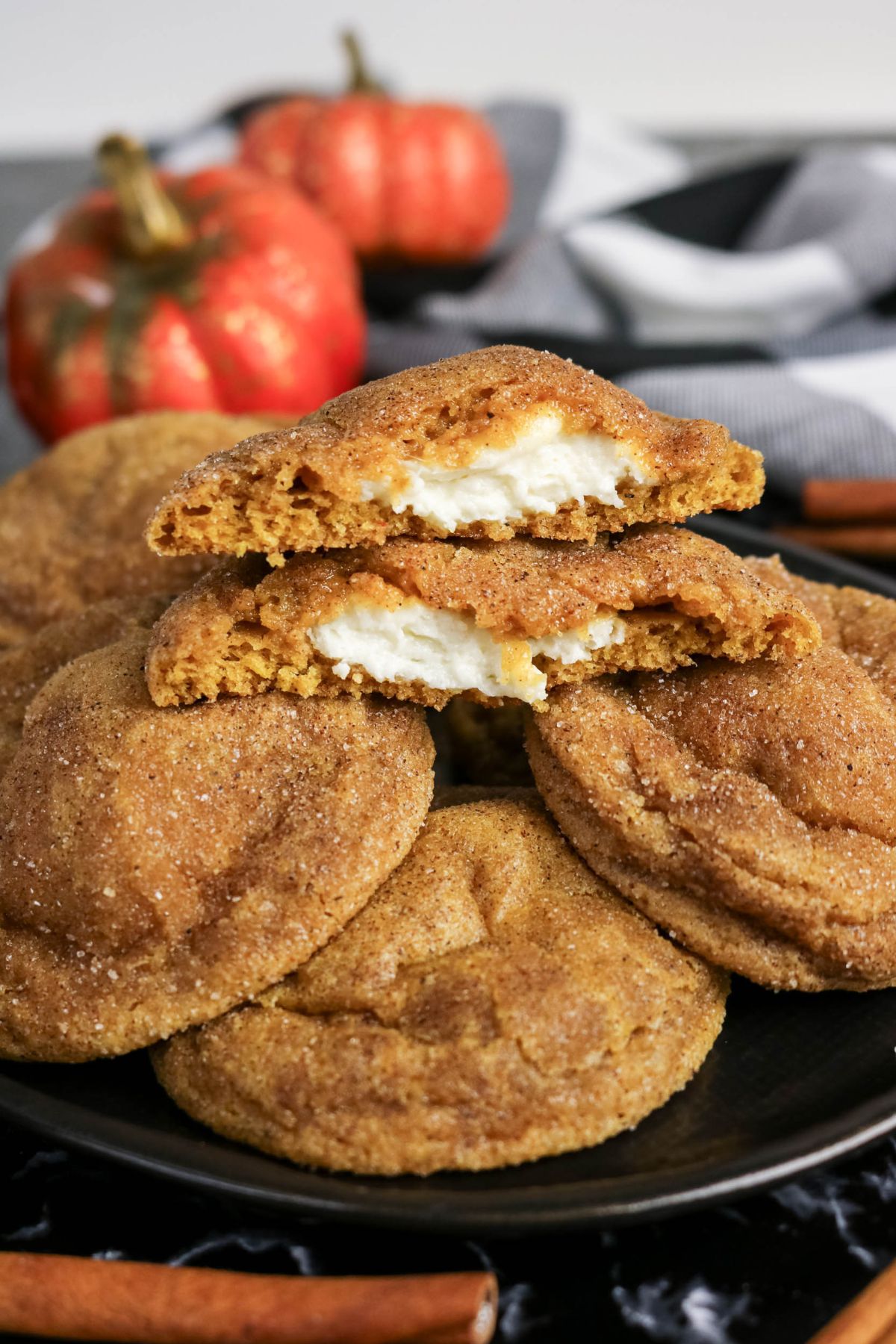 Plate of pumpkin cookies coated in cinnamon sugar, with one cookie broken in half showing a creamy white filling. Two small pumpkins and a cloth are in the background.