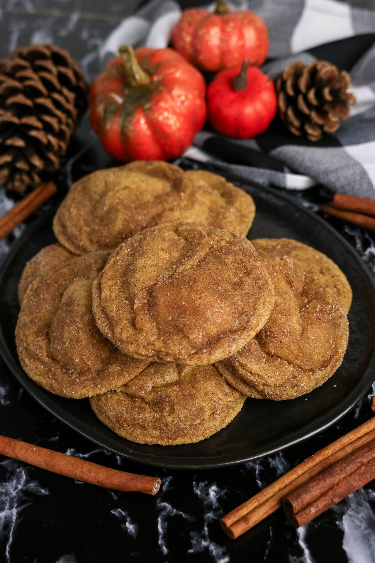 A plate of cinnamon sugar cookies is surrounded by pinecones, small decorative pumpkins, a striped cloth, and cinnamon sticks on a dark marble surface.