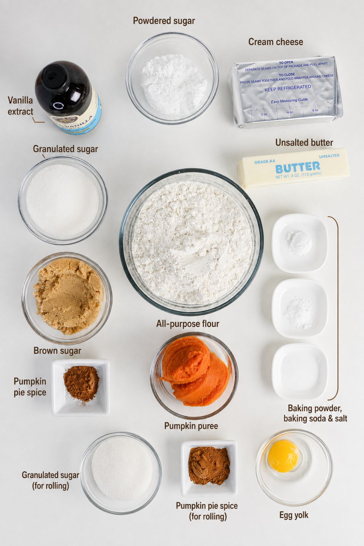 Various labeled ingredients for baking are arranged on a white surface, including sugars, flour, pumpkin puree, egg, butter, cream cheese, vanilla extract, and spices in bowls and containers.