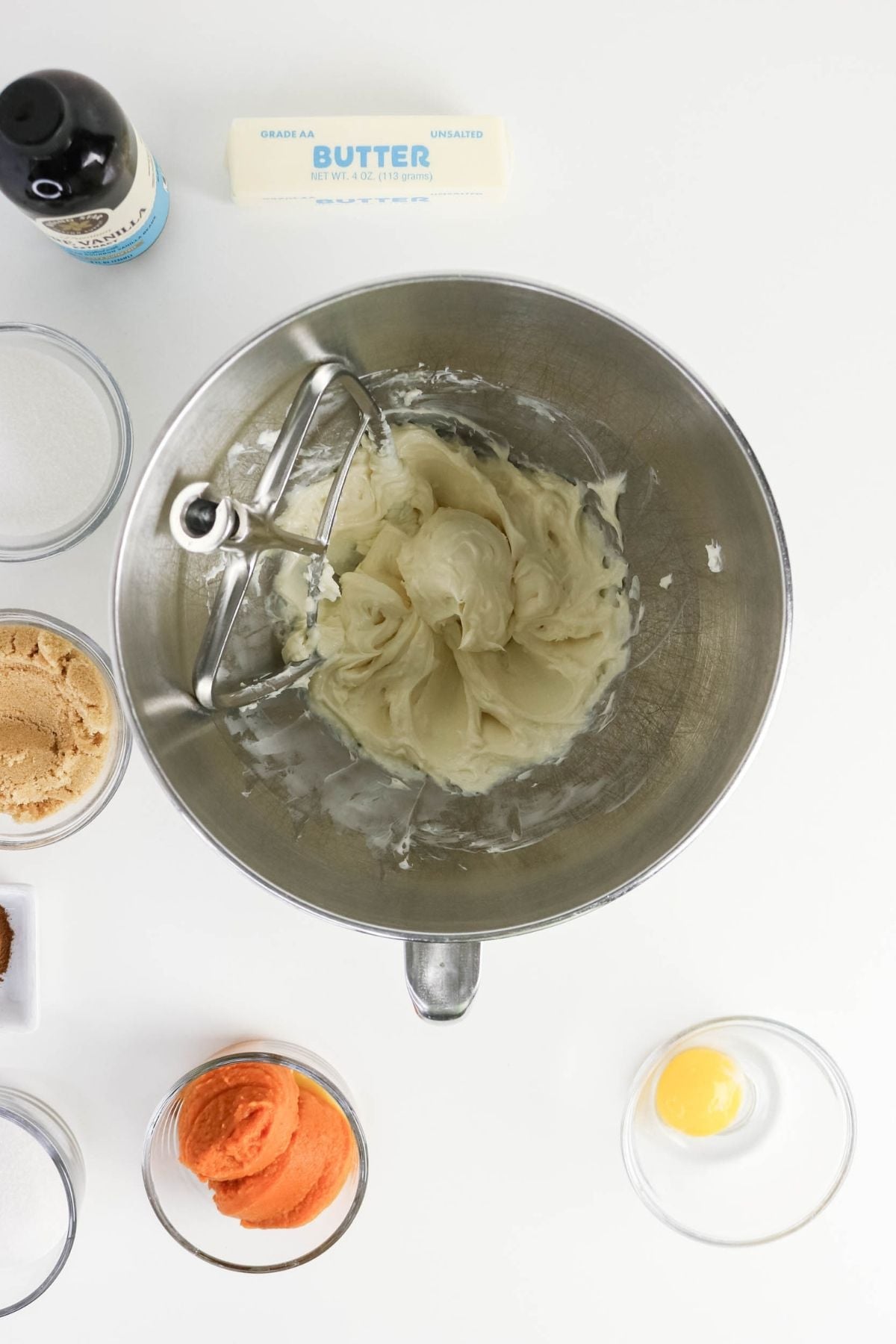 A mixing bowl with creamed butter, surrounded by a stick of butter, vanilla extract, white and brown sugar, pumpkin puree, an egg yolk, and small bowls of other ingredients.