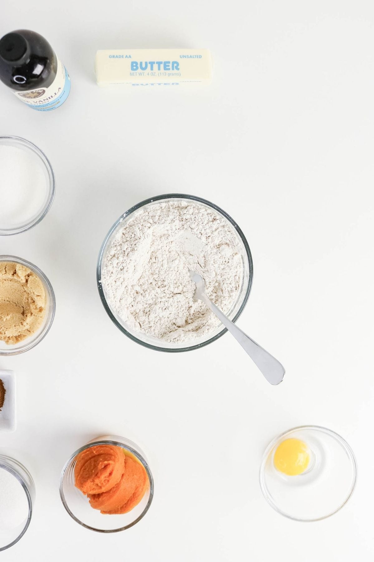 Bowls of flour, sugar, brown sugar, pumpkin puree, an egg, a stick of butter, and a bottle of vanilla are arranged on a white surface, viewed from above.