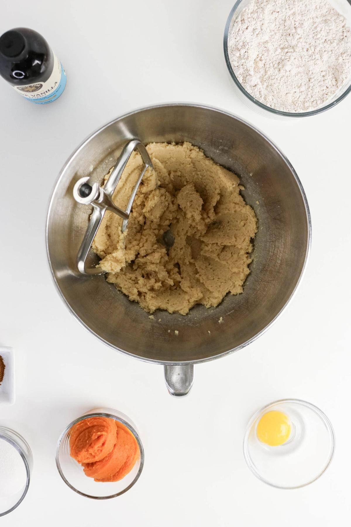 A mixing bowl with creamed butter and sugar, surrounded by a bowl of flour, a bottle of vanilla, pumpkin puree, an egg, and other baking ingredients on a white surface.