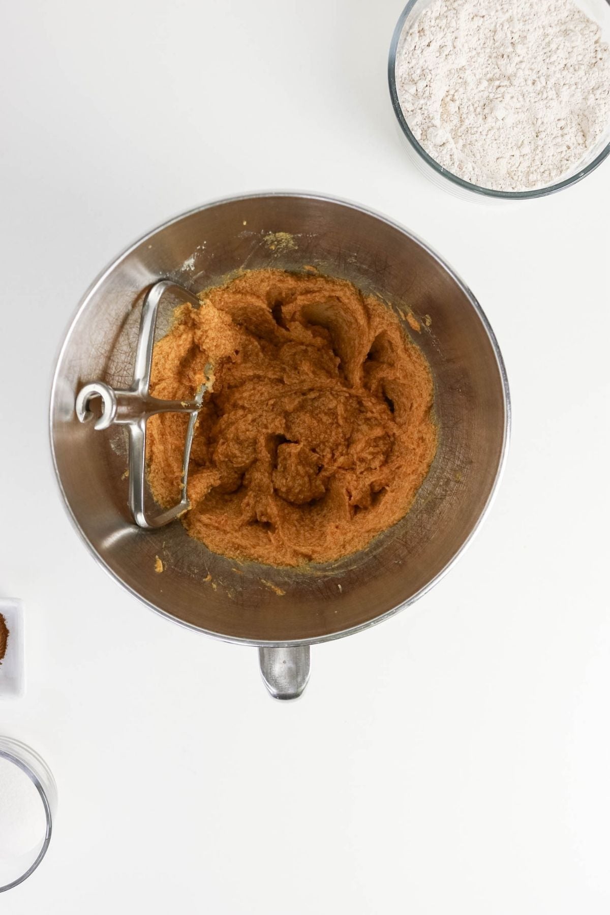 A mixing bowl with brown cookie dough next to bowls of flour, sugar, and spices on a white surface.