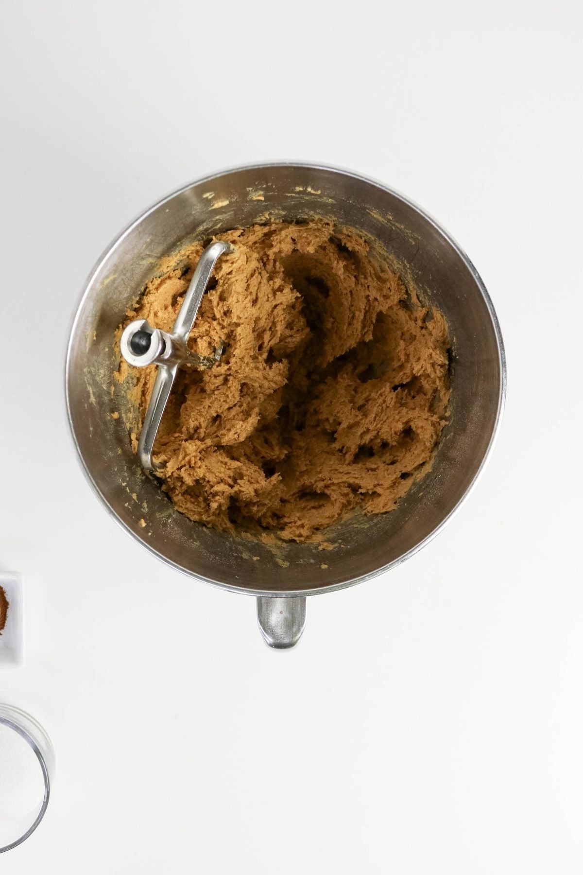 A mixing bowl with a paddle attachment contains brown cookie dough mixture on a white surface.