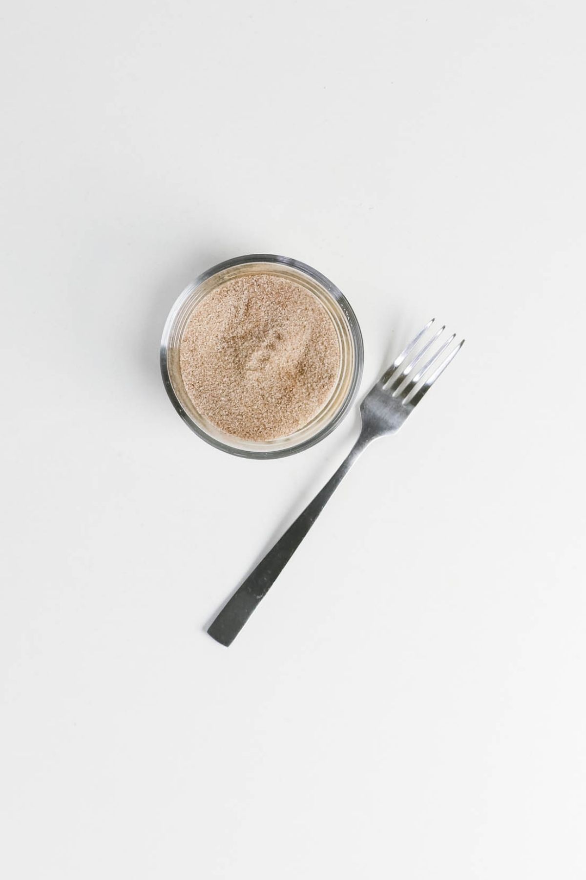 A small glass bowl filled with a light brown powder is next to a metal fork on a white surface.