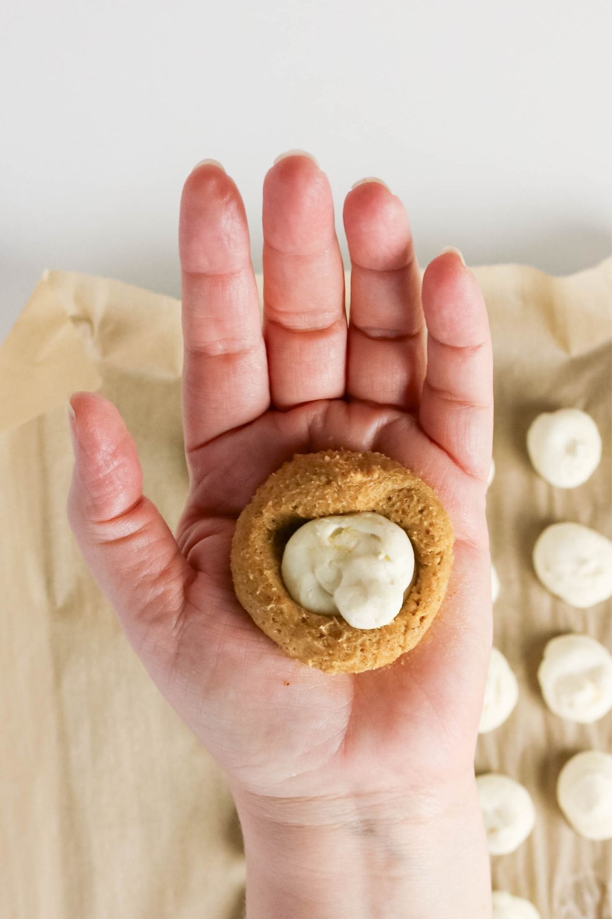 A hand holding a round cookie with a dollop of cream in the center; more cookies on parchment paper in the background.