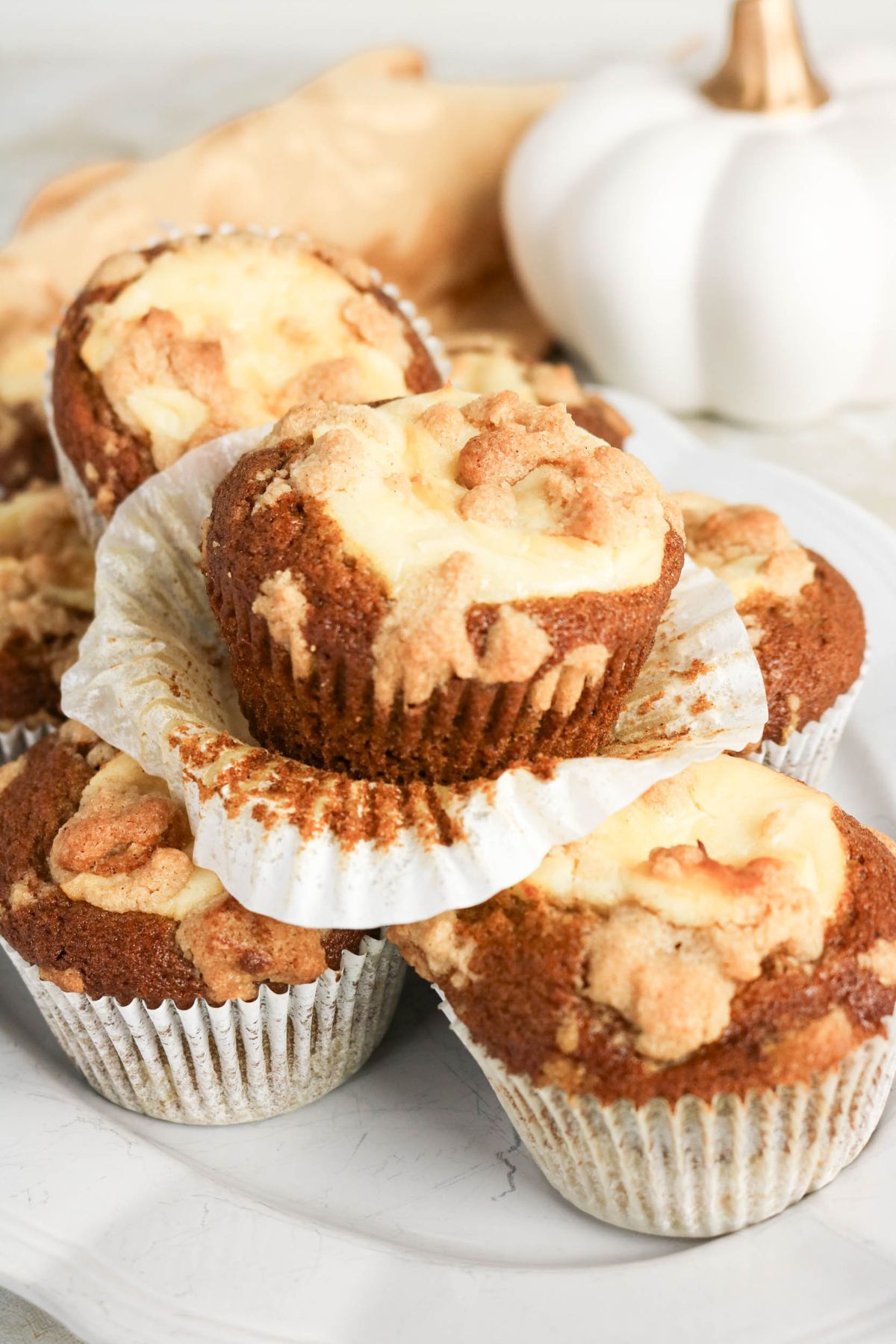 A plate of pumpkin muffins with cream cheese filling and crumb topping, stacked and displayed on a white plate with a white pumpkin decoration in the background.